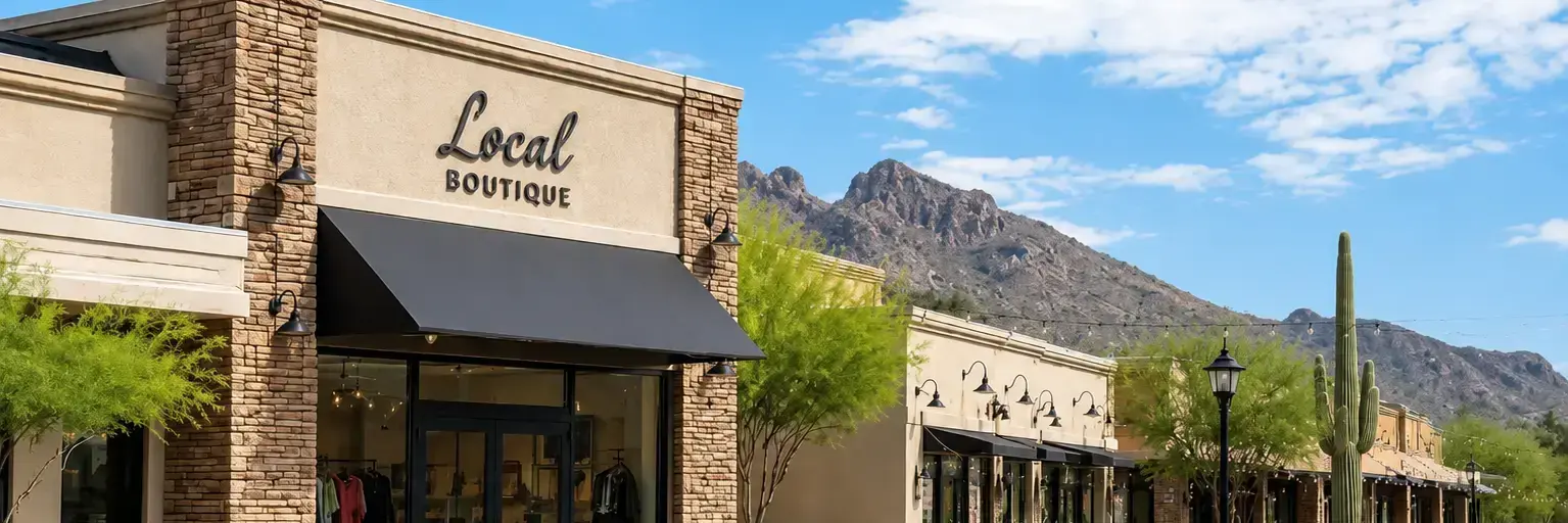 Front of a small shopping plaza with a stone Local Boutique storefront under a black awning, desert landscaping, and mountains in the background.