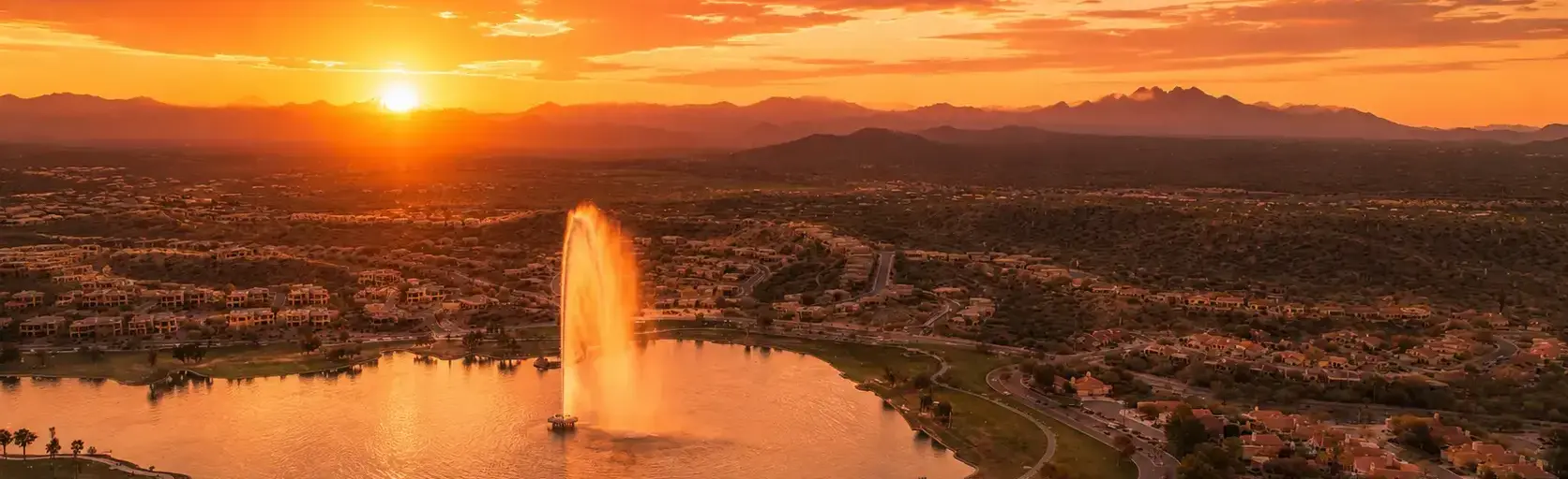Golden hour over a desert town: an orange sunset with a tall water fountain shooting from a circular lake, surrounded by houses and winding roads.