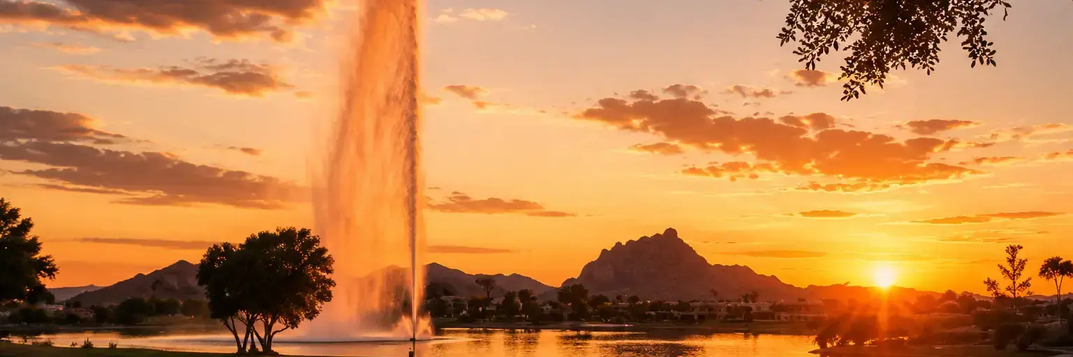 Sunset over a lake with a tall water fountain, trees and mountains silhouetted in the distance.